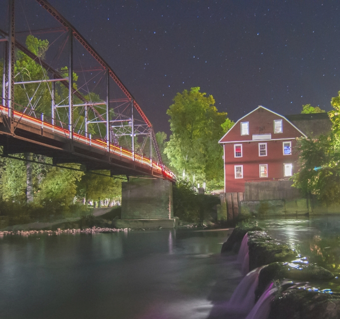 red barn next to a river with a lit bridge at night